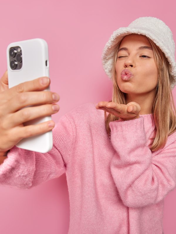 Romantic young woman sends air kiss at cellular camera takes flirty selfie photo winks eye and pouts lips wears hat and jumper isolated over pink background enjoys video chat with boyfriend.